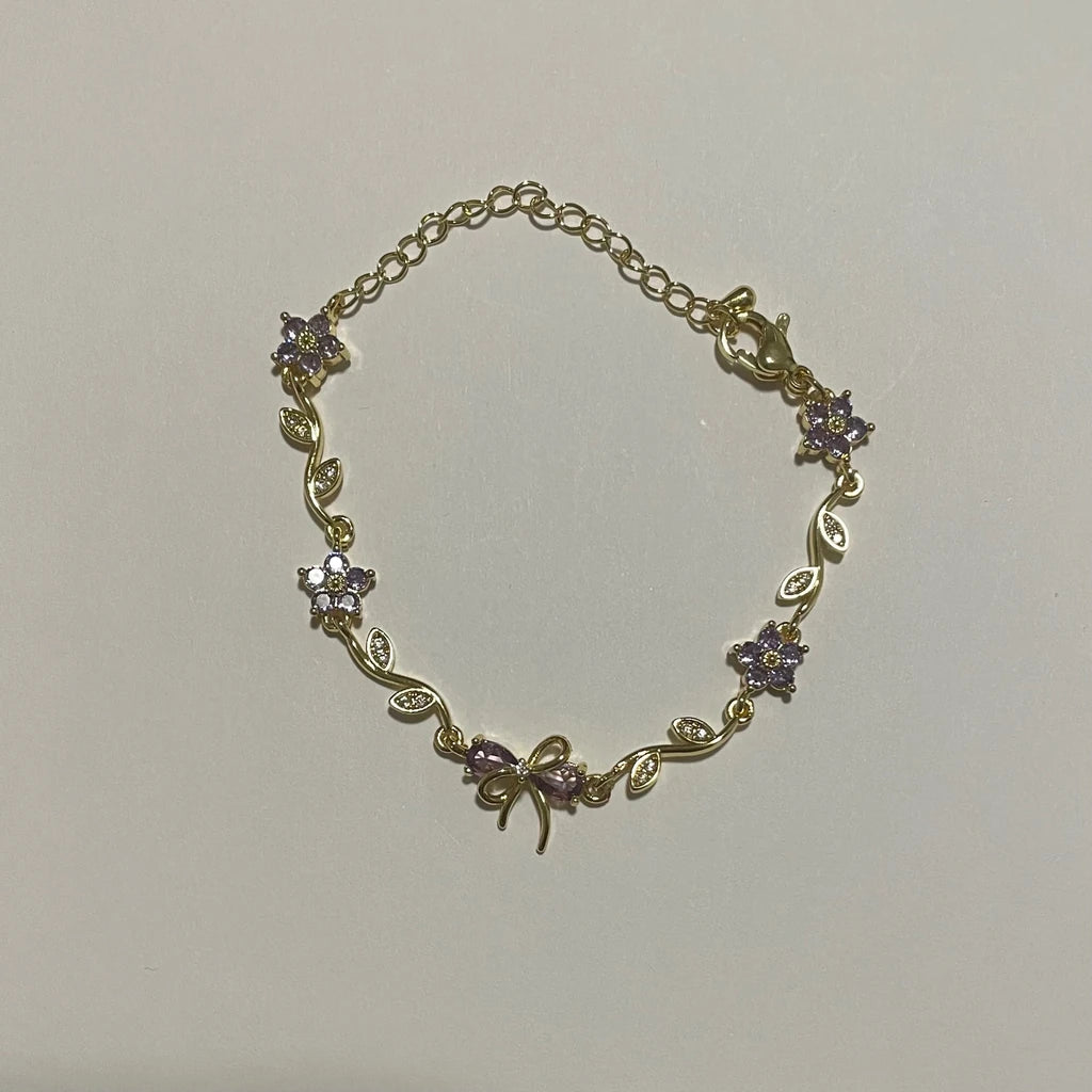Bracelet de couleur dorée à fleurs violettes et motif nœud, avec fermoir mousqueton et chaînette de rallonge