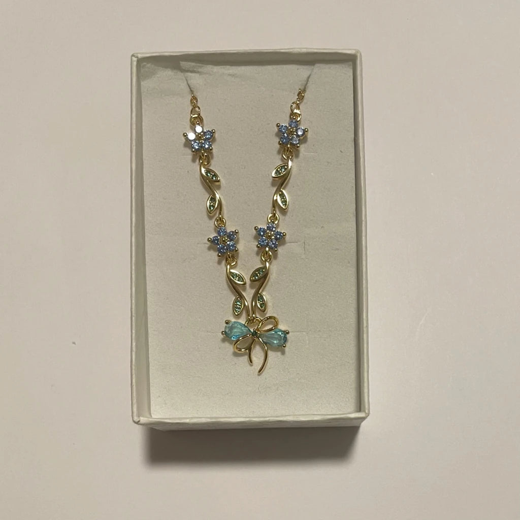 Collier doré à fleurs bleues, feuilles vertes et nœud turquoise, présenté dans un écrin clai