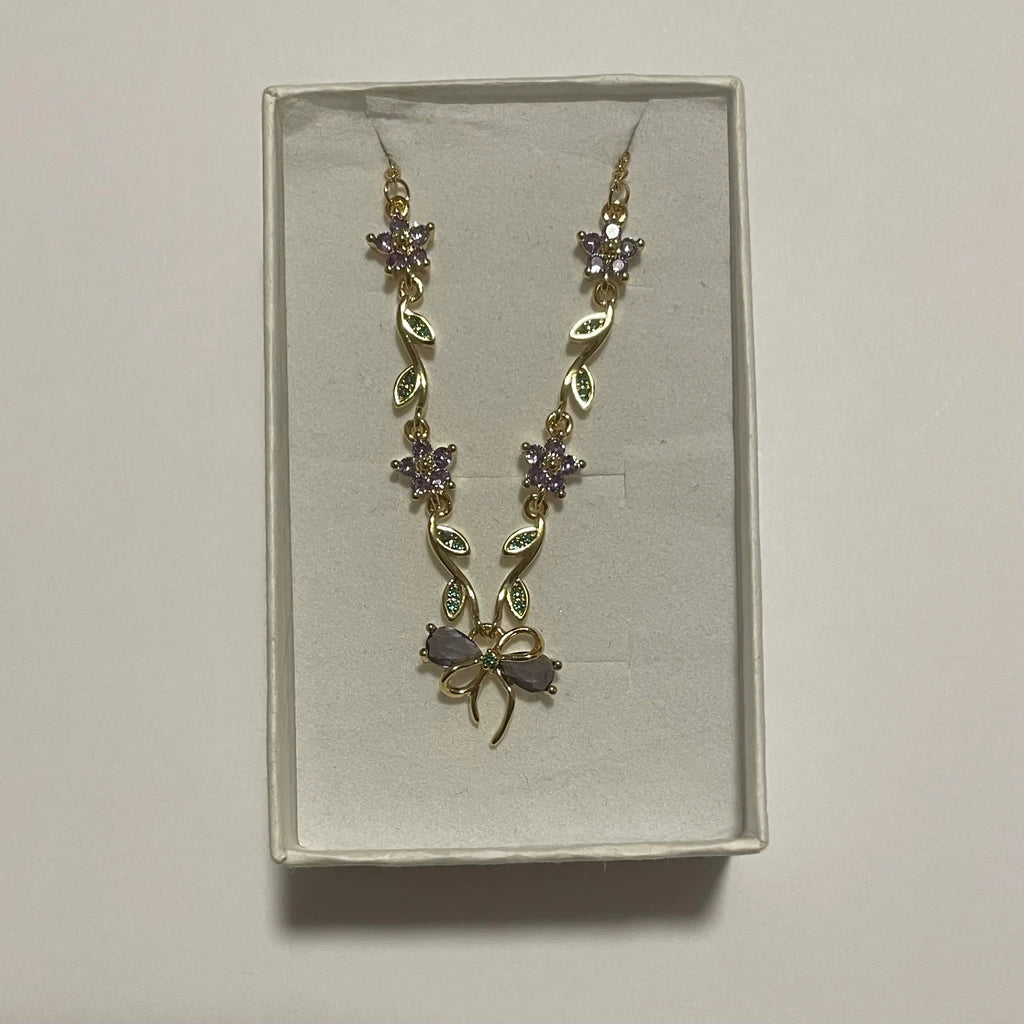 Collier de couleur dorée à fleurs violettes et motif nœud, présenté dans un écrin clair